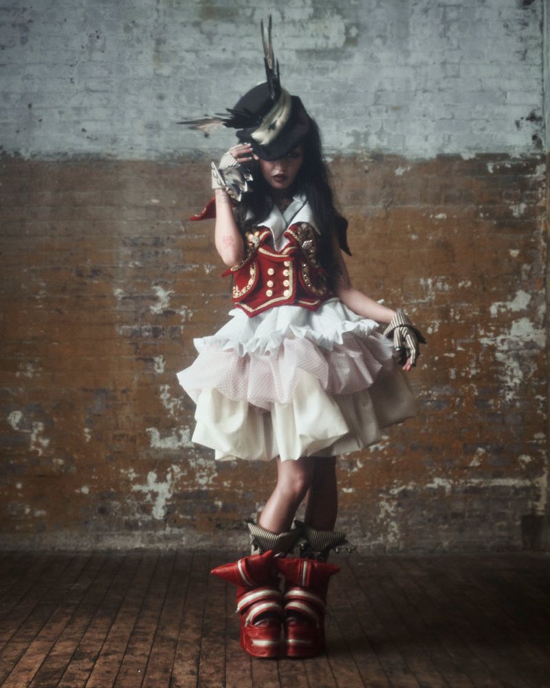 Woman in theatrical costume and elaborate headdress poses against a distressed brick wall with aged wooden flooring in a studio setting.