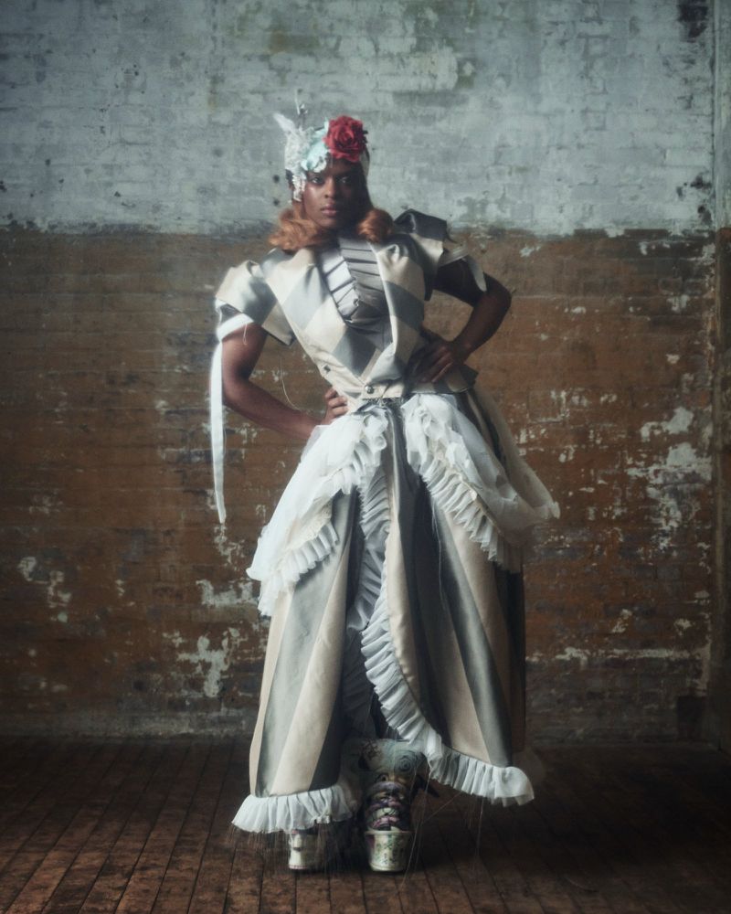 Model standing in a textured gown with ruffles, stripes, and a floral headpiece against a weathered, two-toned brick wall.