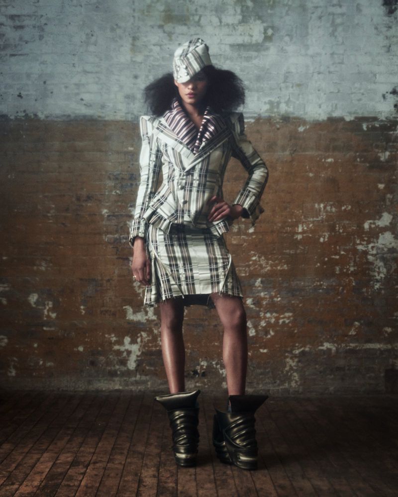 A person stands in a studio wearing a plaid matching skirt, jacket, and hat with large black boots, in a fashion editorial image.