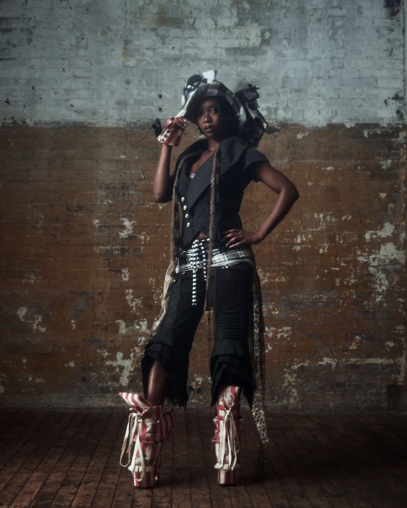 A stylish Black woman stands with hand on hip in unique fashion ensemble with hat and platform boots against a brick studio wall.