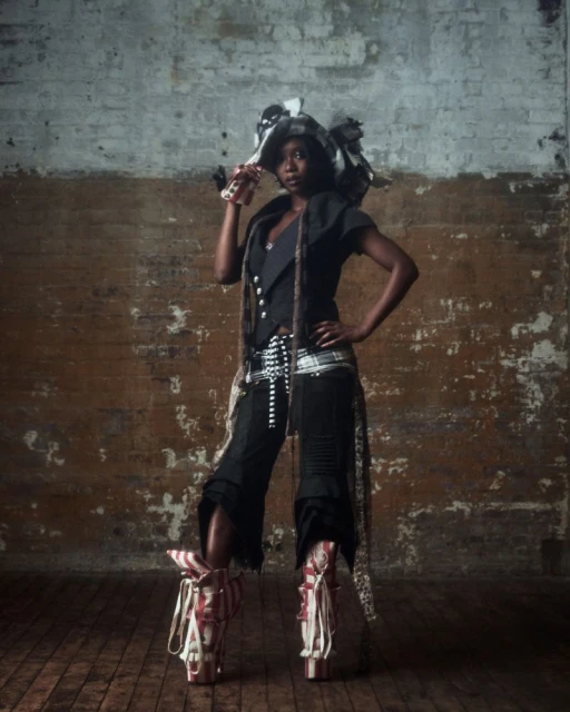 A stylish Black woman stands with hand on hip in unique fashion ensemble with hat and platform boots against a brick studio wall.