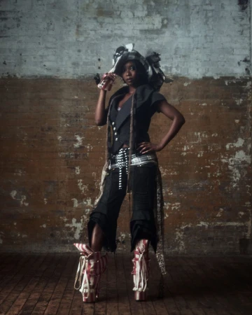 A stylish Black woman stands with hand on hip in unique fashion ensemble with hat and platform boots against a brick studio wall.