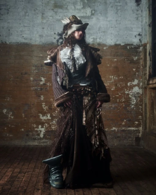 Person in ornate brown coat, frilled shirt, and feathered hat poses indoors against distressed brick wall and wooden floor.