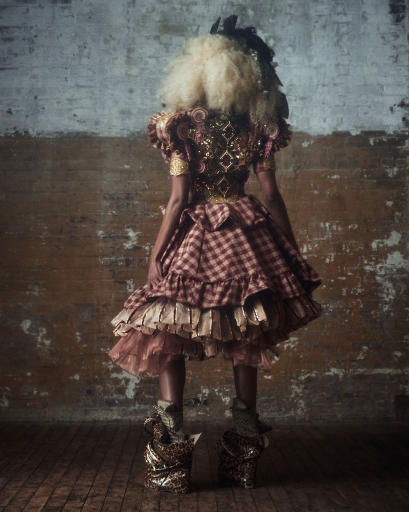 Person in an elaborate, ruffled costume with red plaid, gold accents, and a blonde wig, stands against a textured brick wall.
