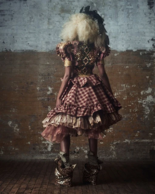 Person in an elaborate, ruffled costume with red plaid, gold accents, and a blonde wig, stands against a textured brick wall.