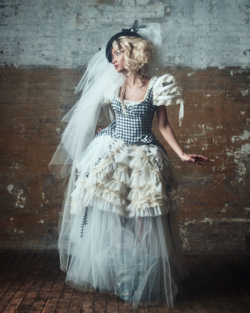 A woman with blonde wavy hair wearing a black and white gingham corset dress with a layered tulle skirt and a veil, standing against a brick wall.