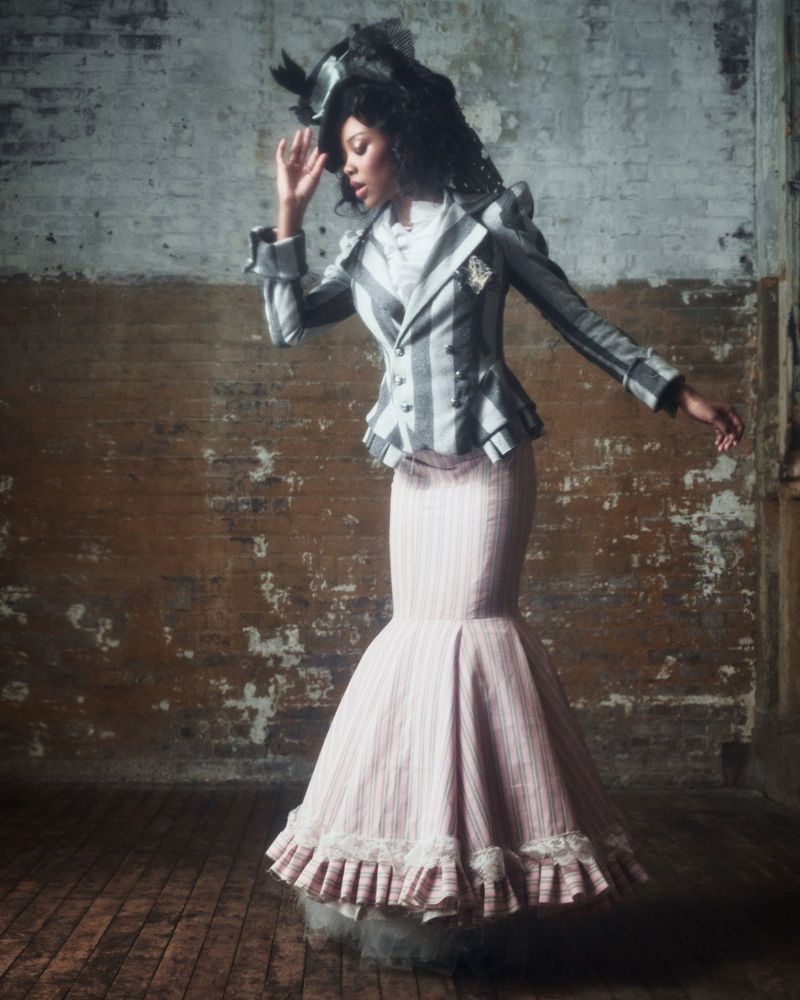 Fashionable woman in striped jacket and skirt poses indoors. Victorian-inspired design elements and vintage brick wall add character.