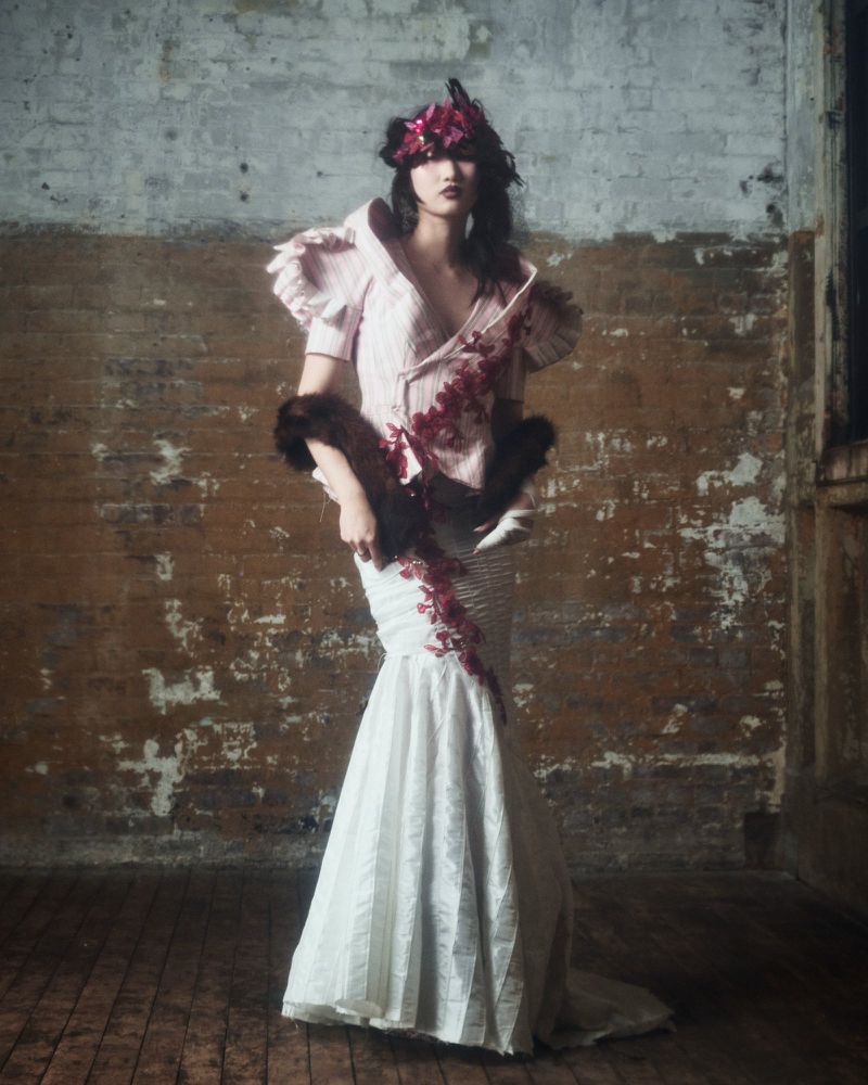 A full shot of a model in an ornate white mermaid dress and floral headpiece against a worn brick wall, creating a dramatic fashion portrait.