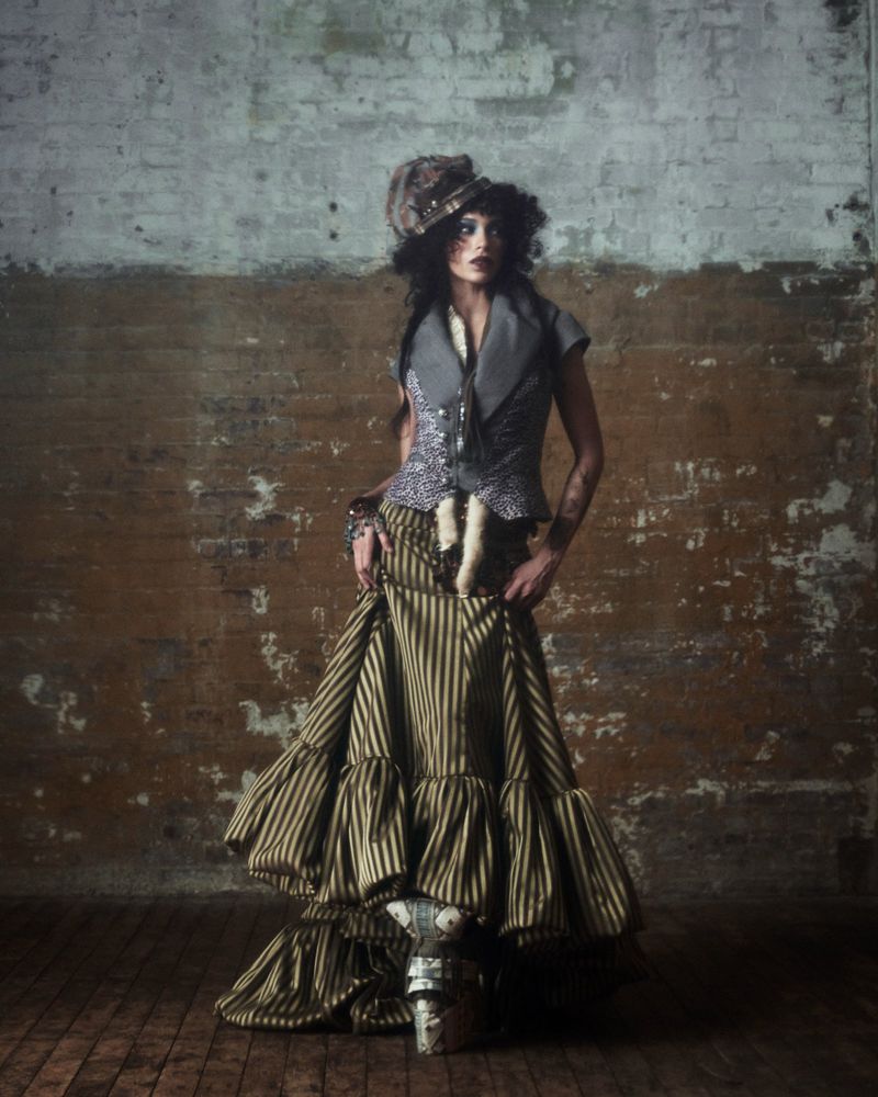 A woman stands in a striped dress and ornate hat against a distressed brick wall, fashion portrait, studio lighting, full shot.