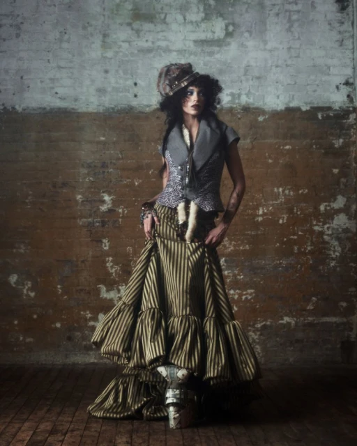 A woman stands in a striped dress and ornate hat against a distressed brick wall, fashion portrait, studio lighting, full shot.