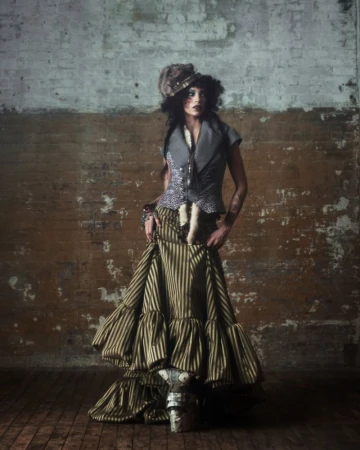 A woman stands in a striped dress and ornate hat against a distressed brick wall, fashion portrait, studio lighting, full shot.