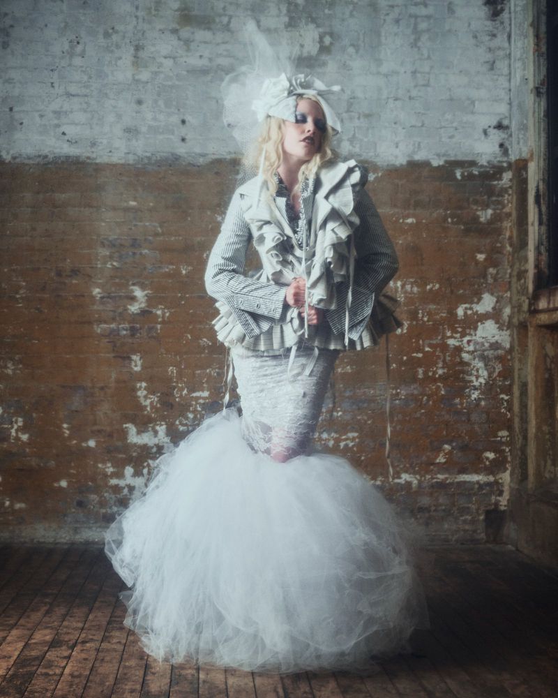 A woman in haute couture fashion poses in a textured silver and white ruffled jacket and puffy skirt against a weathered brick wall.