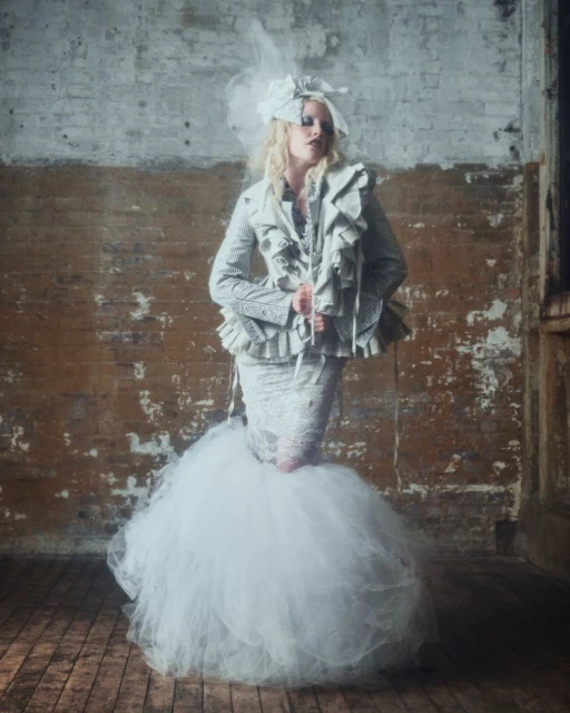 A woman in haute couture fashion poses in a textured silver and white ruffled jacket and puffy skirt against a weathered brick wall.