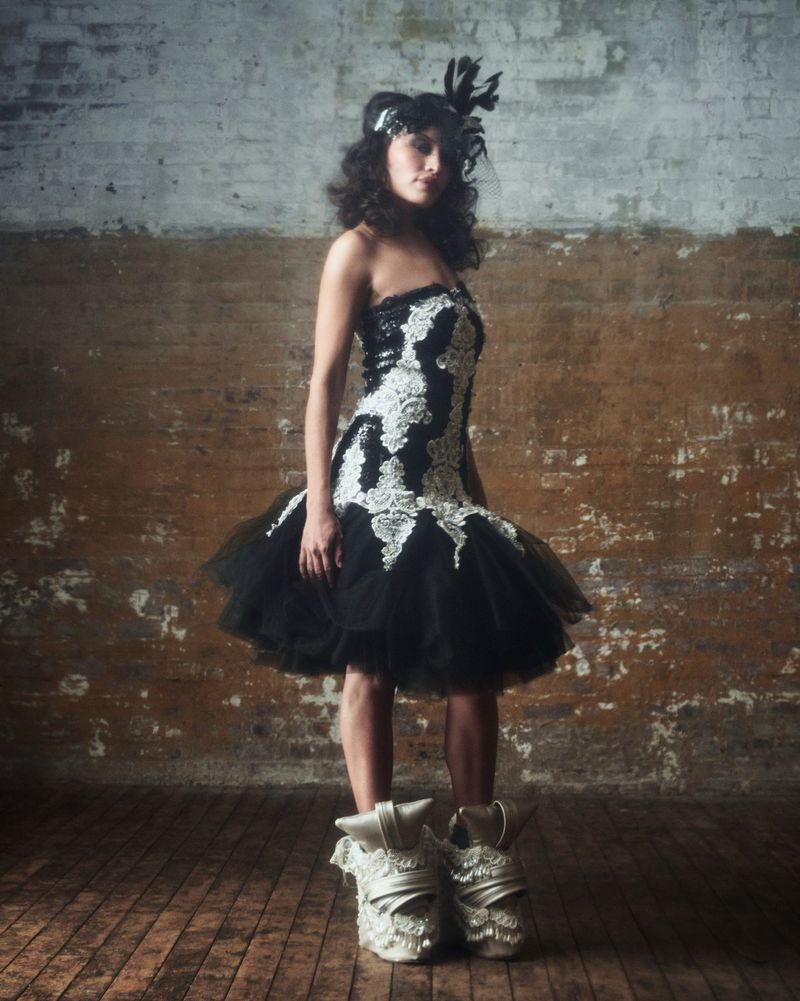 Full-length portrait of an adult woman posing in a black and white tutu dress with feathered headpiece and ornate high-heeled shoes in studio.