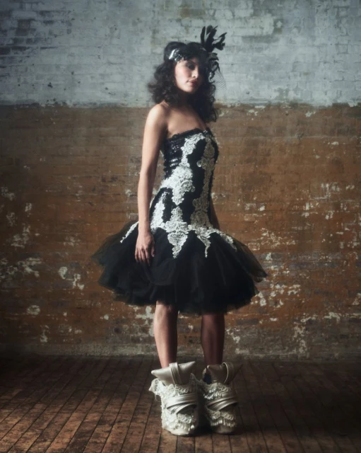 Full-length portrait of an adult woman posing in a black and white tutu dress with feathered headpiece and ornate high-heeled shoes in studio.