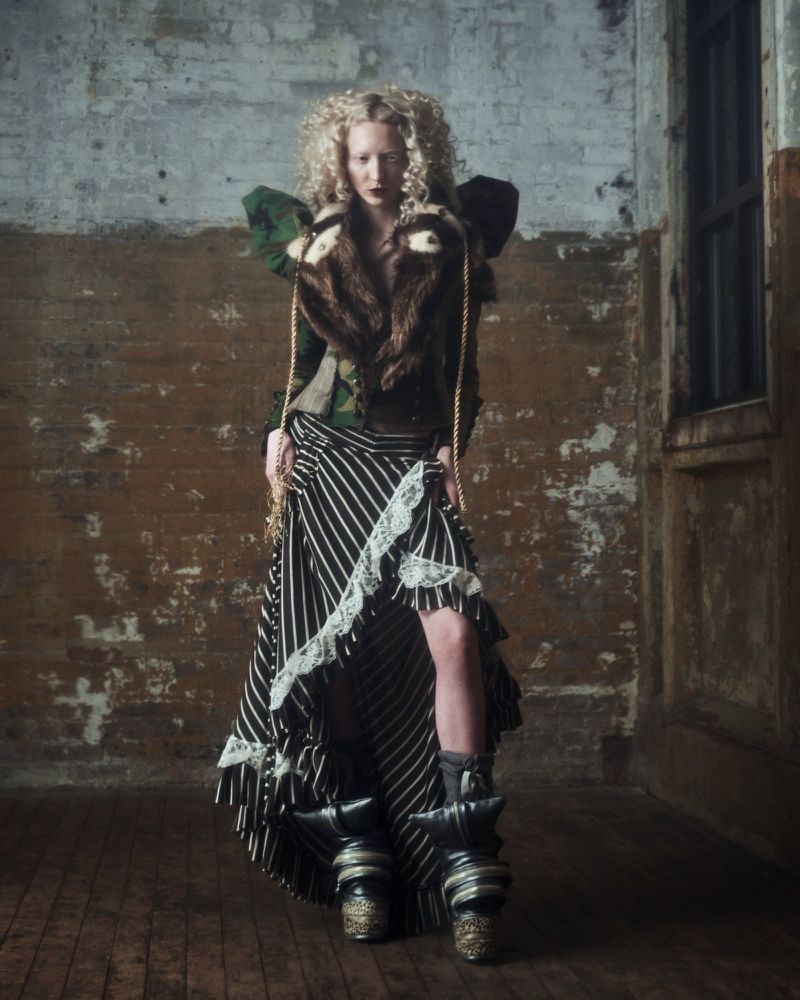 A fair-skinned, curly-haired woman in an avant-garde outfit stands in a distressed interior wearing a brown fur-collared jacket and striped skirt.