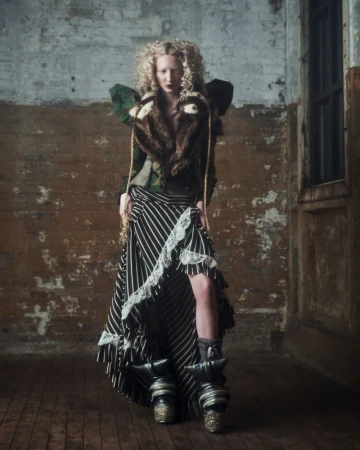 A fair-skinned, curly-haired woman in an avant-garde outfit stands in a distressed interior wearing a brown fur-collared jacket and striped skirt.