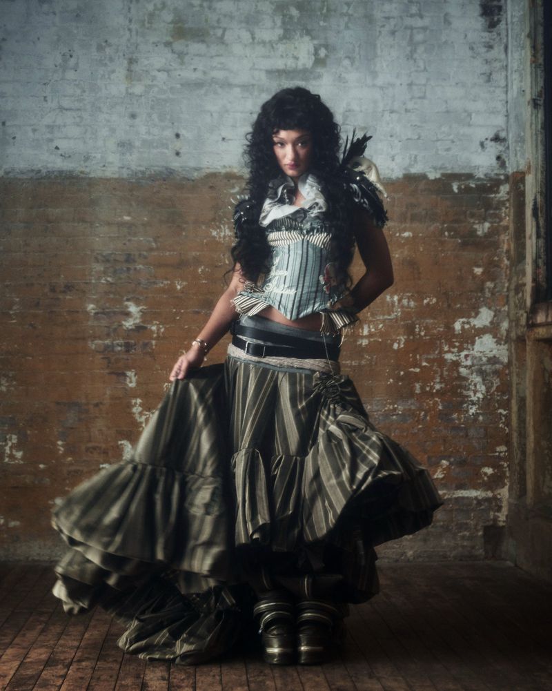 Woman poses in vintage striped fashion with corset, layered skirt, and Victorian-style boots indoors against a distressed brick wall.