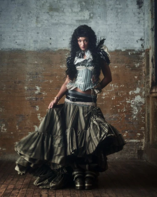 Woman poses in vintage striped fashion with corset, layered skirt, and Victorian-style boots indoors against a distressed brick wall.