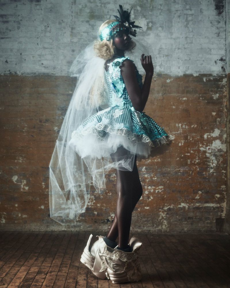 A striking image of a dark-skinned woman wearing a blue-and-white dress with feather accents and avant-garde shoes, posing in a studio.