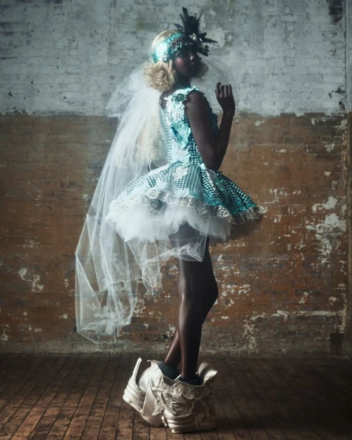 A striking image of a dark-skinned woman wearing a blue-and-white dress with feather accents and avant-garde shoes, posing in a studio.