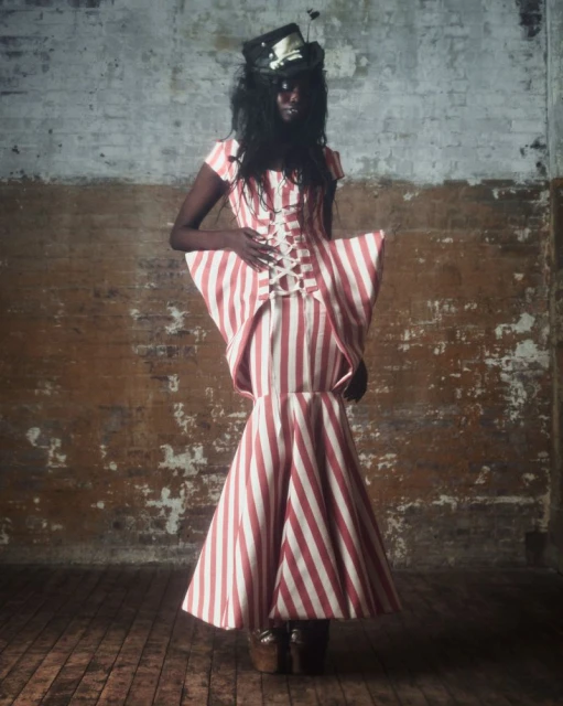 Adult female fashion model poses in a red-and-white-striped gown with a black top hat against a distressed wall in a studio.