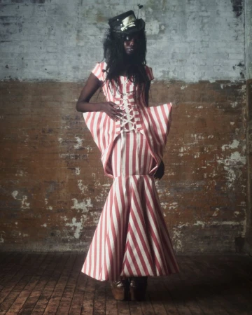Adult female fashion model poses in a red-and-white-striped gown with a black top hat against a distressed wall in a studio.