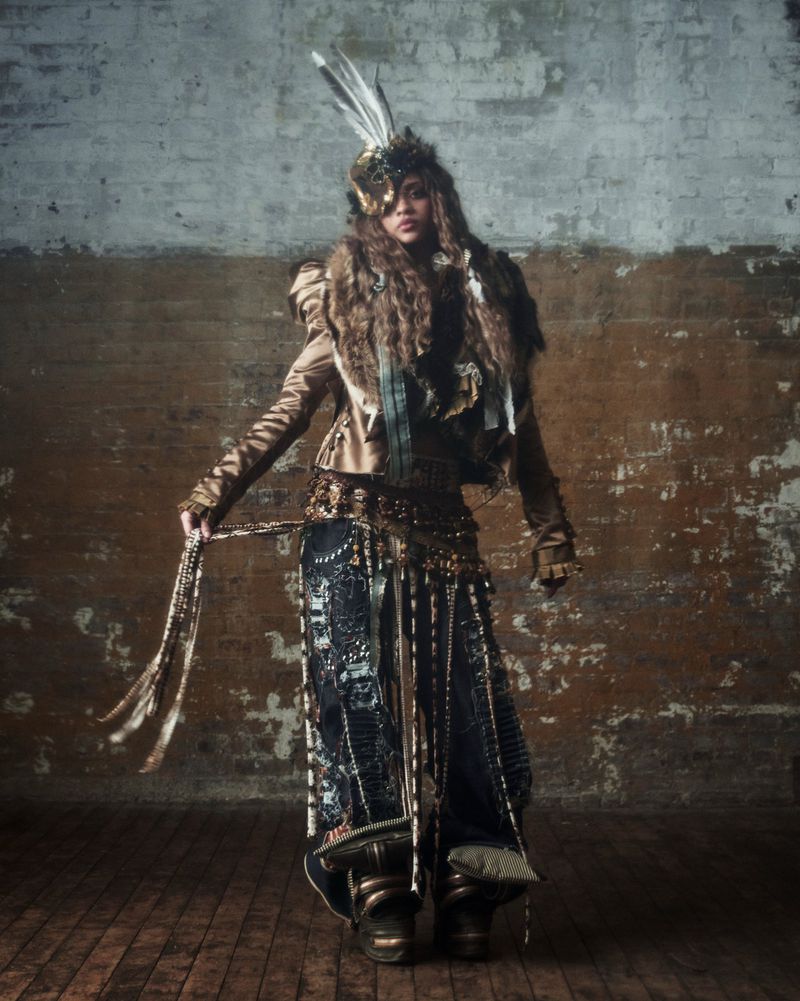 Full-length studio shot of an adult female model in a steampunk-inspired outfit with feathered headpiece, jacket, and skirt against a textured wall.