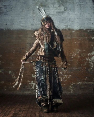 Full-length studio shot of an adult female model in a steampunk-inspired outfit with feathered headpiece, jacket, and skirt against a textured wall.