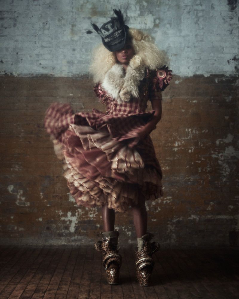 Model in a plaid dress with flared skirt and feathered hat standing against a distressed brick wall interior