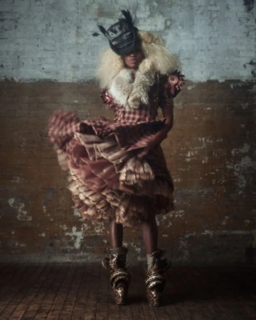 Model in a plaid dress with flared skirt and feathered hat standing against a distressed brick wall interior