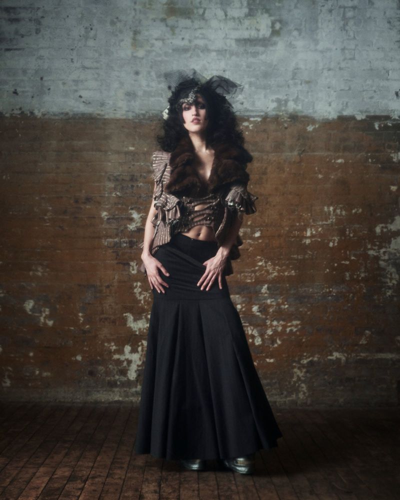 Elegant woman in a fashion-forward outfit with a fur stole and long black skirt poses against a weathered brick wall in a studio shot.