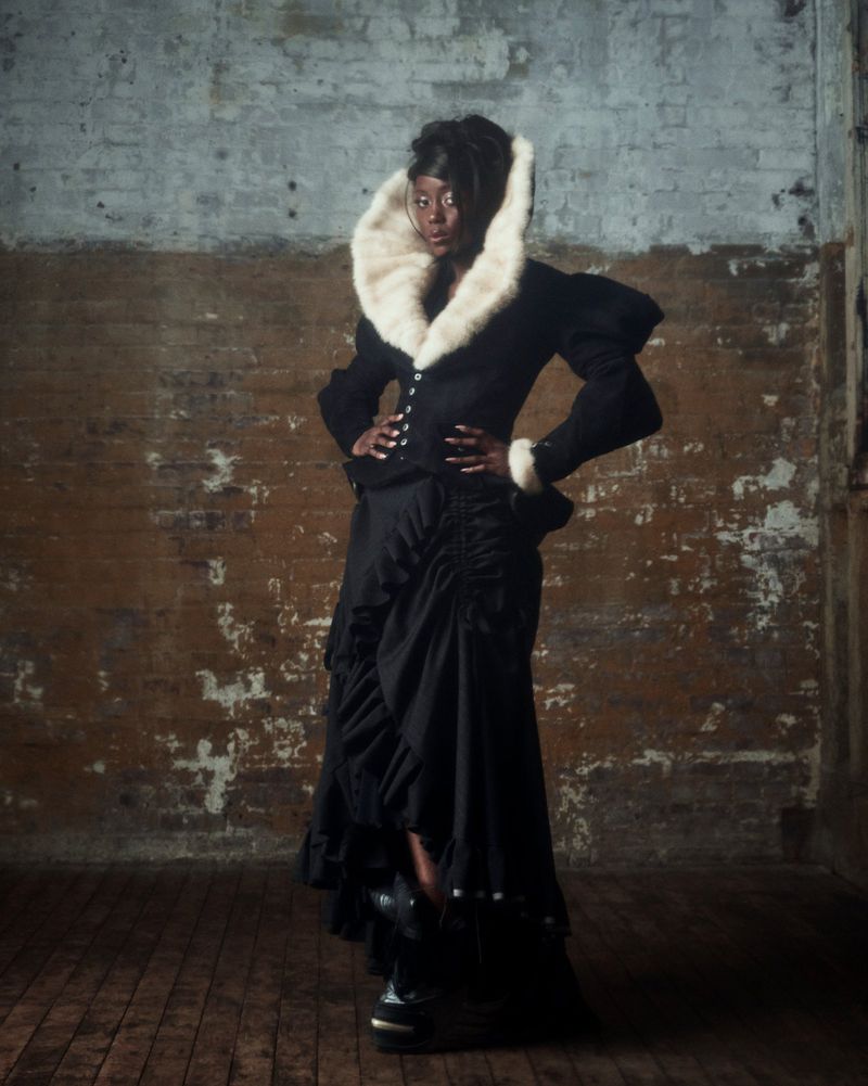 A confident Black woman poses in a stylish black outfit with a striking white fur collar against a textured brick wall in a studio setting.
