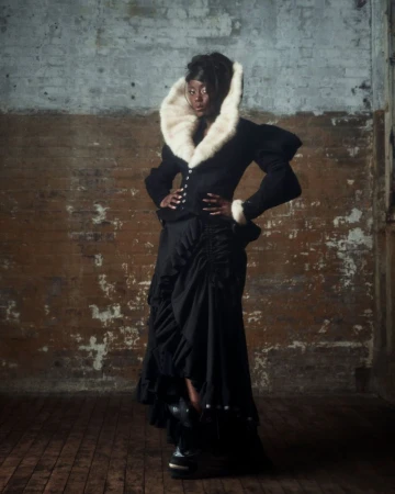 A confident Black woman poses in a stylish black outfit with a striking white fur collar against a textured brick wall in a studio setting.