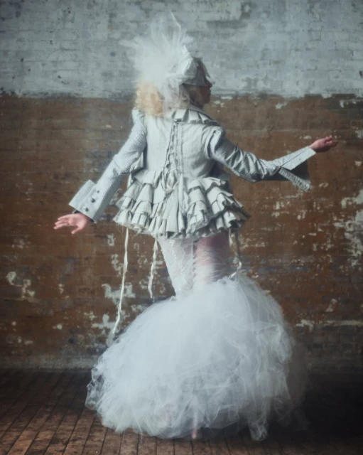 Female in white gown, textured jacket and feathered headwear poses indoors against a textured wall and wood floor in soft lighting.