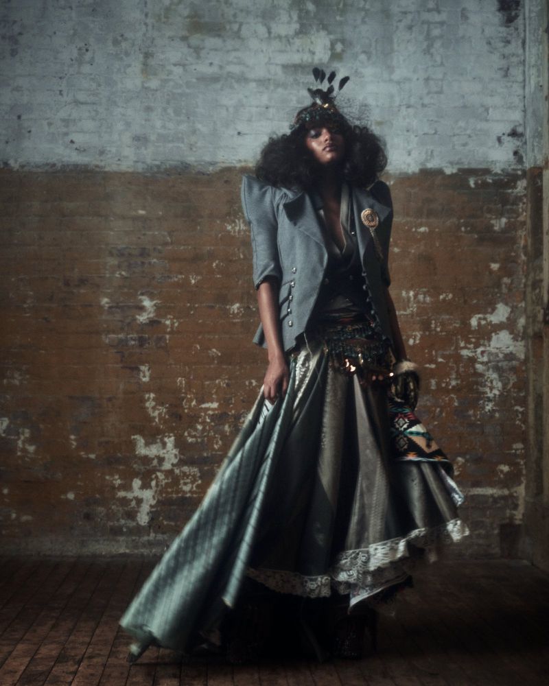 A woman with an afro poses in a gray jacket and long skirt in a photo studio with a distressed brick wall and wood floor.