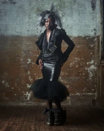 Model in avant-garde black structured jacket and sequined skirt with sheer tulle headpiece standing before a weathered brick wall.