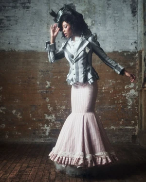 Model in a striped Victorian jacket and ruffled skirt posing against an exposed brick wall interior