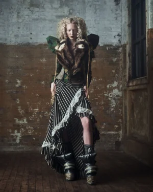 Model with blonde curly hair wearing a patterned jacket and striped skirt standing in an industrial room