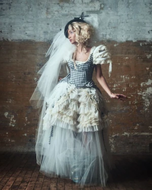 Model in a voluminous layered white tulle dress and checkered corset with a veil against a distressed brick wall.