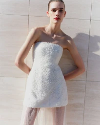 Model wearing a strapless white textured mini dress posing against a stone tile wall for Simkhai Endless Summer 2025 campaign.