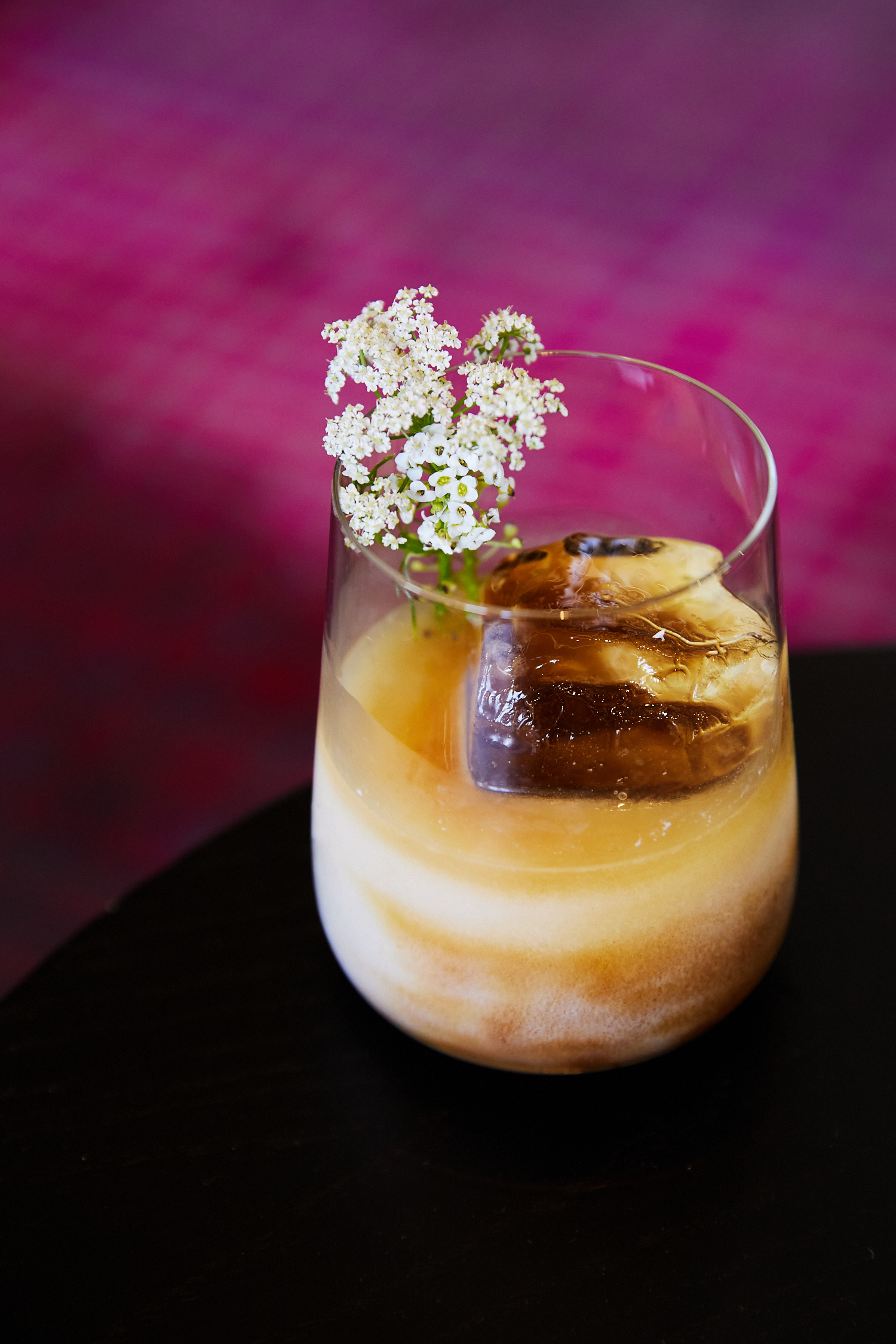 Cocktail in a glass with an ice cube, garnished with white flowers, set against a textured magenta background, on a black surface.