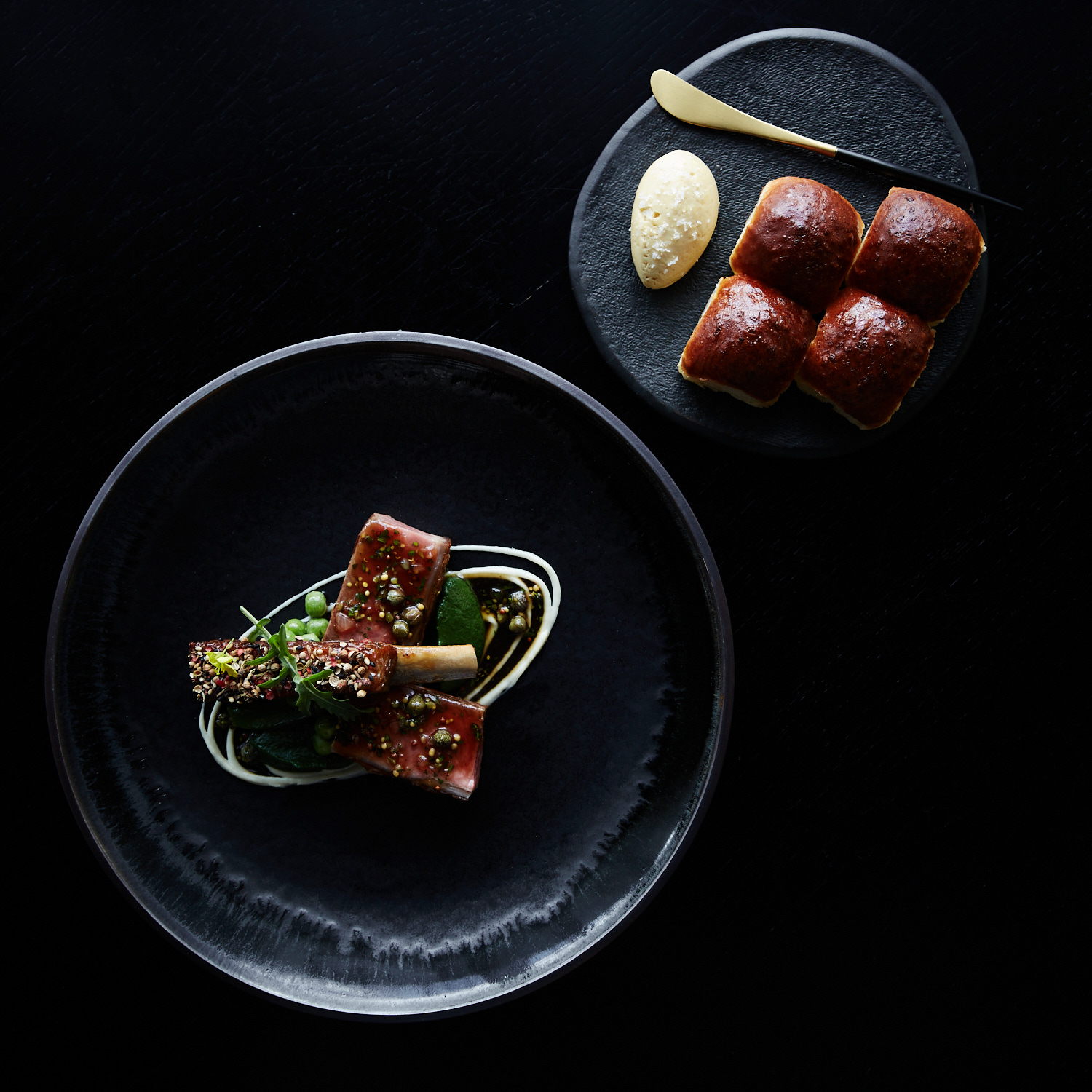 Overhead shot of plated lamb dish with vegetables and sauce on a black plate, alongside dinner rolls and butter on a dark surface.
