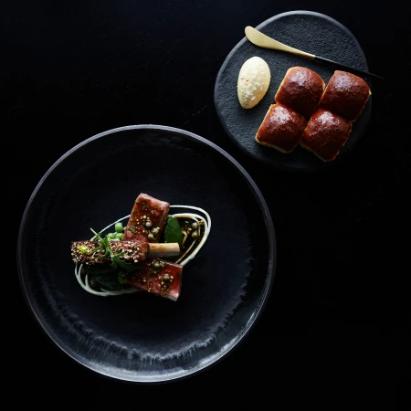 Overhead shot of plated lamb dish with vegetables and sauce on a black plate, alongside dinner rolls and butter on a dark surface.