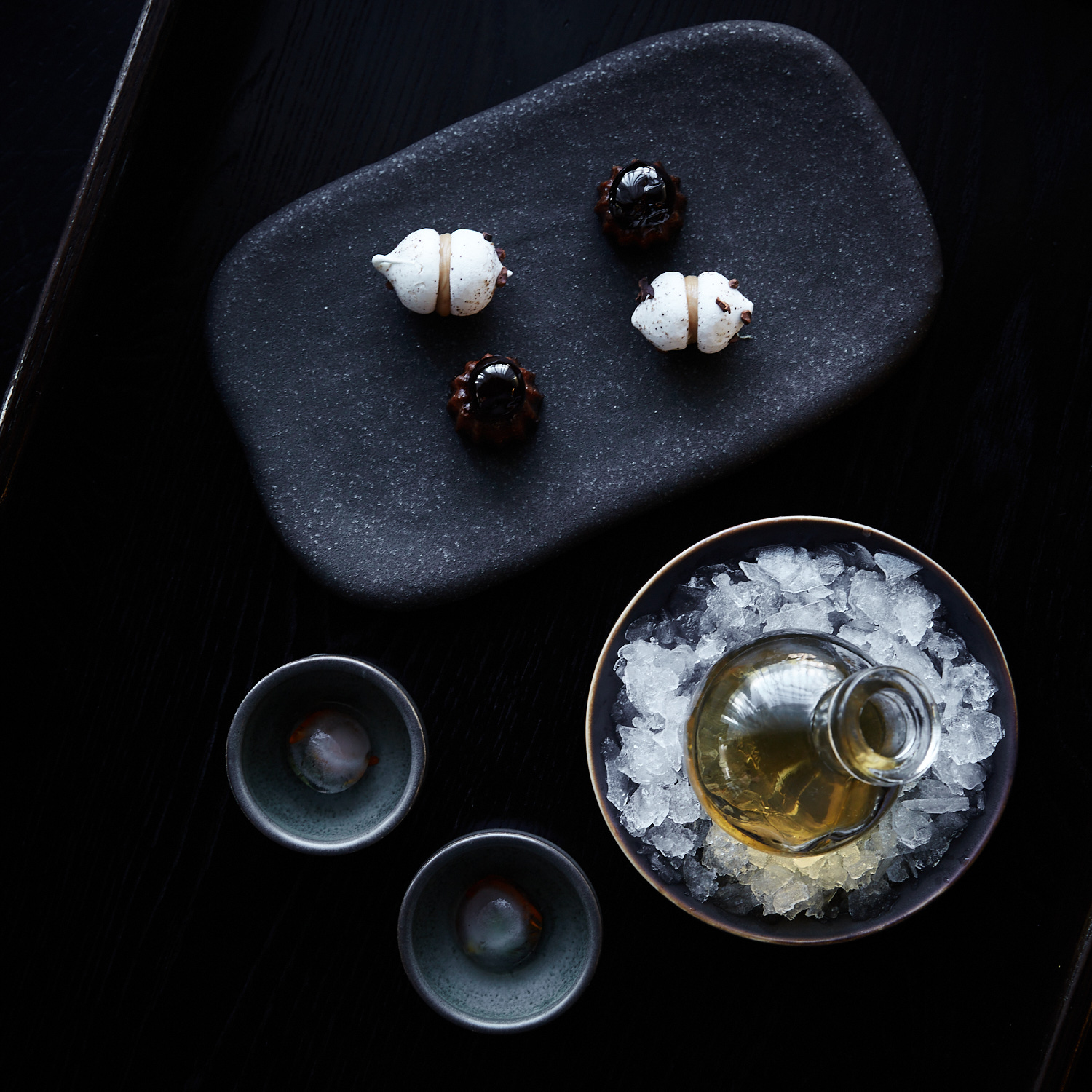Overhead shot of dark tabletop with sake bottle surrounded by ice, sake cups, and small desserts on a rectangular plate.