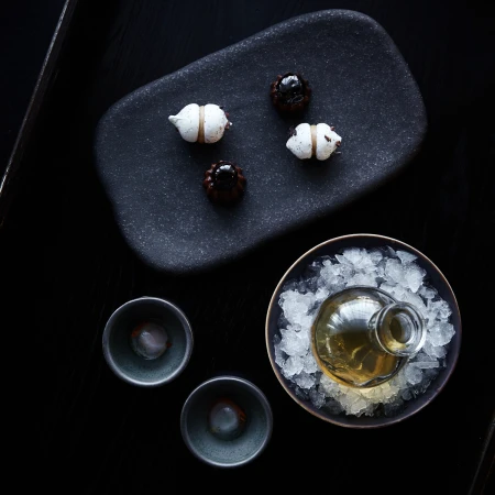Overhead shot of dark tabletop with sake bottle surrounded by ice, sake cups, and small desserts on a rectangular plate.