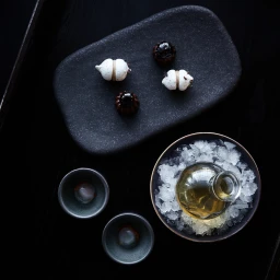 Overhead food photograph of small pastries, two small ceramic bowls, and a glass bottle nested in ice on a black surface