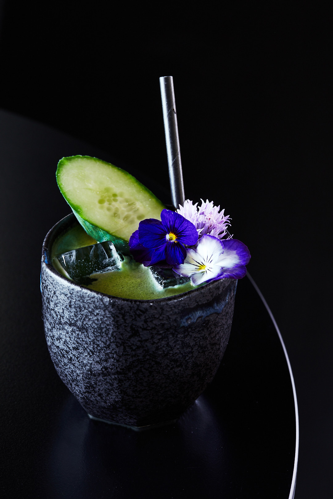 Cocktail in a dark grey speckled glass, garnished with a cucumber slice, purple and white flowers, and a metal straw on a black surface.