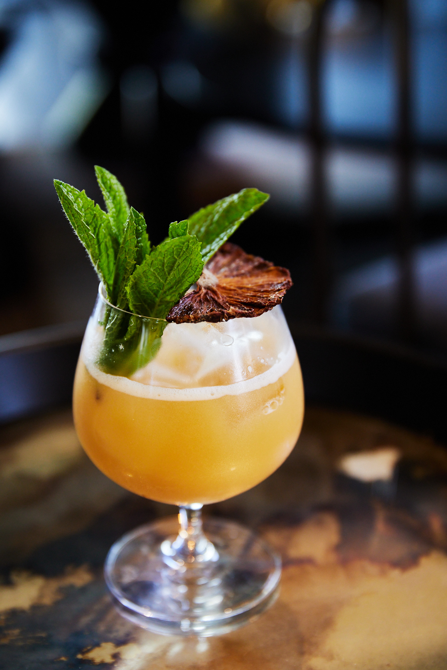 A stem glass filled with an orange-colored cocktail, garnished with fresh mint leaves and a dehydrated orange slice, on a reflective surface.
