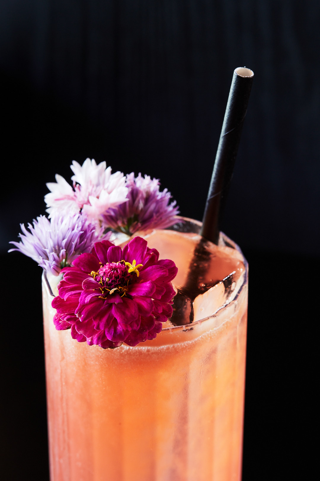 A close-up shot of a refreshing cocktail in a tall glass, garnished with vibrant pink and purple flowers and a black straw.